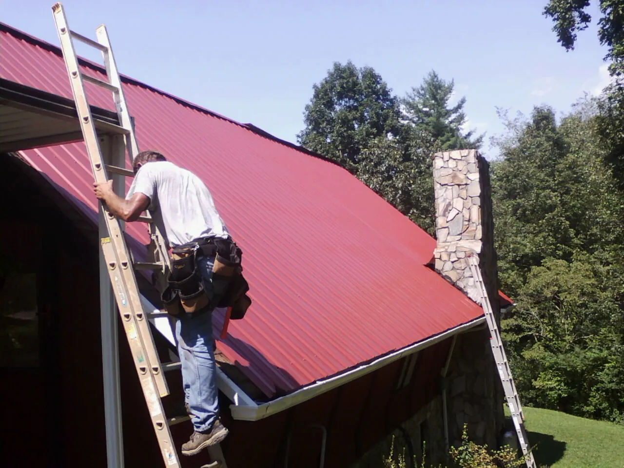 Roofer on a ladder beside a red metal roof and stone chimney — Soffit & Fascia Repair by Roofing Brothers in Salyer